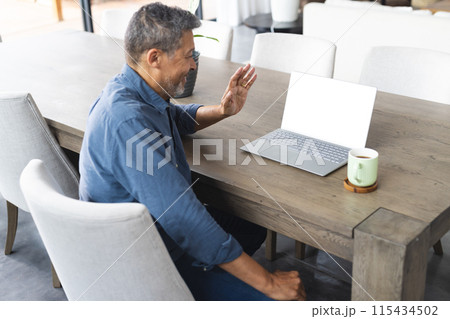Biracial senior man in a blue shirt waves during a video call on his laptop 115434502