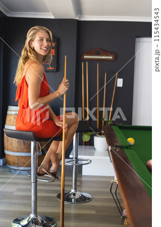 Young Caucasian woman in an orange dress enjoys a game of pool, holding a cue stick with copy space Young Caucasian woman in an orange dress enjoys a game of pool, holding a cue stick with copy space 115434543