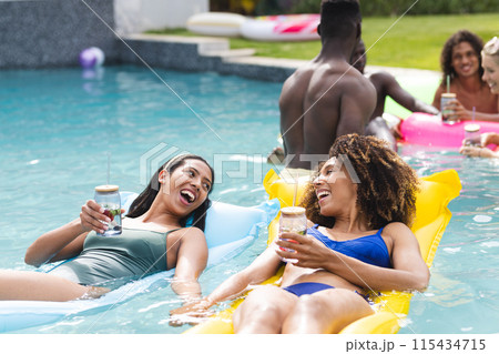 Diverse group enjoys a sunny pool party, with two women laughing on floaties 115434715