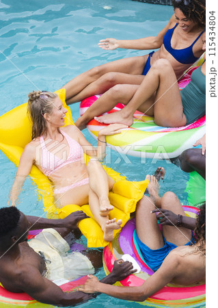 Diverse group of friends enjoy a pool party, lounging on colorful floaties 115434804