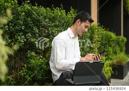 Millennial businessman using mobile phone while sitting outdoors cafe surrounded by green plants 115436924