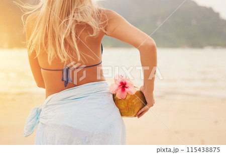 Bikini, woman and coconut cocktail at beach on vacation, travel and summer holiday on island in Greece. Back view, fruit drink and girl at sea to relax in nature with healthy organic hibiscus flower 115438875