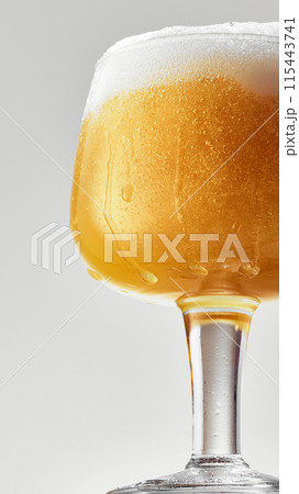 Close-up of mug filled with golden, hazy beer topped with thick, frothy white foam against grey studio background. Close-up of mug filled with golden, hazy beer topped with thick, frothy white foam against grey studio background. 115443741