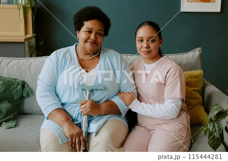 Young nurse carefully holding mature African American womans hand while they sitting on sofa and posing for photo Young nurse carefully holding mature African American womans hand while they sitting on sofa and posing for photo 115444281