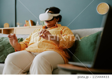 Mature African American woman sitting on sofa and getting in touch with virtual reality 115444433