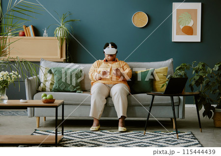 African American elderly woman sitting on sofa and interacting with VR game 115444439