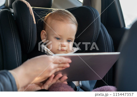 Mother is giving a tablet to her toddler daughter sitting in car seat to entertain her during long road trip Mother is giving a tablet to her toddler daughter sitting in car seat to entertain her during long road trip 115447357
