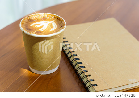 Hot coffee latte with latte art milk foam in cup made of paper and notebook on wood desk on top view. As breakfast In a coffee shop at the cafe,during business work concept 115447529
