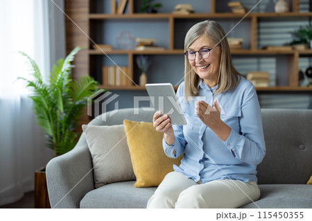 Senior woman having a video call using a tablet while sitting on a sofa at home, smiling and enjoying the moment. 115450355