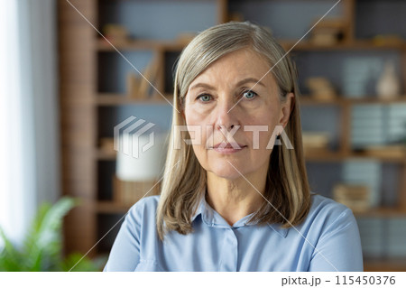 A professional portrait of a senior Caucasian businesswoman in a blue shirt, looking directly at the camera with a confident and calm expression in a well-lit office environment. 115450376