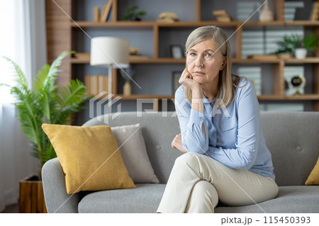 A thoughtful looking mature woman sitting in her cozy living room, gazing into the distance. The peaceful home environment reflects a moment of solitude or contemplation. 115450393