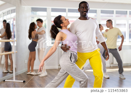 Smiling young woman wearing sportswear dancing in couple samba dance with african american partner during group class at dance salon 115453863