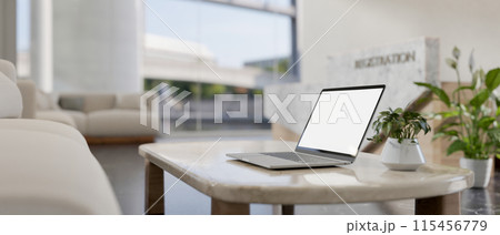 A laptop computer with a white-screen mockup on a coffee table in a modern reception lounge area. 115456779