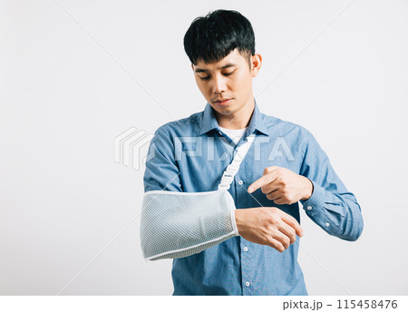 Portrait Asian smiling young man wearing a cast on his arm broken studio shot isolated white background, men using splint for treatment pointing finger to his arm, Concept of discomfort and pain 115458476