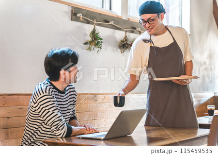コーヒーを配膳するカフェの店員・ウェイター・オーナーの男性とテレワークする男性客 115459535