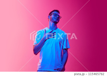 Pleasure. Portrait of young man in glasses and white t-shirt holding hand on chest and expressing positive vibe against pink background in neon light Pleasure. Portrait of young man in glasses and white t-shirt holding hand on chest and expressing positive vibe against pink background in neon light 115465782