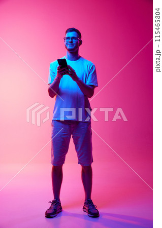 Full-length image of young man in glasses and casual clothes using mobile phone for communication against pink background in neon light 115465804
