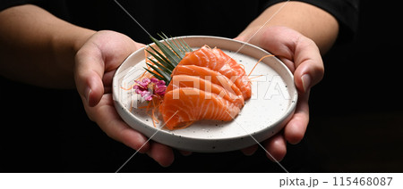 Chef holding ceramic plate with salmon sashimi on black background. Japanese food style concept Chef holding ceramic plate with salmon sashimi on black background. Japanese food style concept 115468087