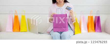 Happiness young asian woman sitting on sofa open paper bag while online shopping with credit card. Happiness young asian woman sitting on sofa open paper bag while online shopping with credit card. 115476293