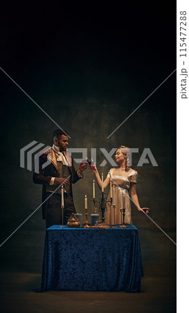 Elegant couple in vintage attire drinking wine over candlelit table against dark, moody background. Romantic evening Elegant couple in vintage attire drinking wine over candlelit table against dark, moody background. Romantic evening 115477288