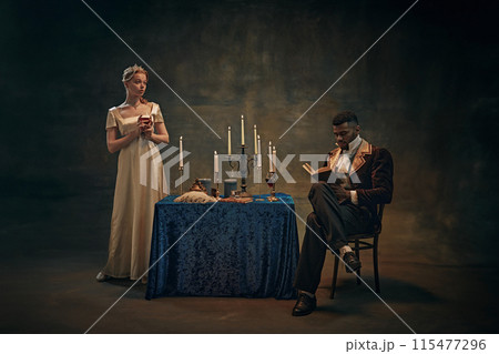 Woman in satin dress holds wine glass, standing beside candlelit table, while man in vintage attire reading book against retro tapestry background 115477296