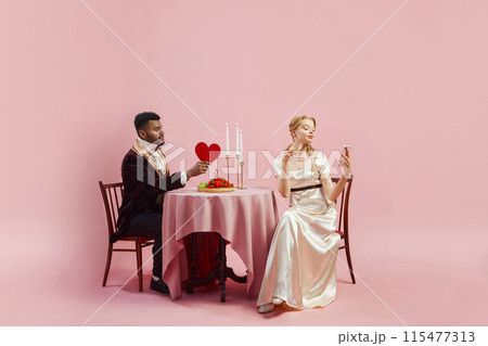 Victorian Valentine. Gentleman shares Valentine heart with lady in classic white gown looking on phone against pink background. 115477313