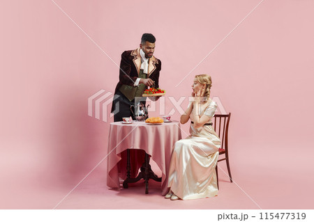 Man in rich velvet jacket offers tray of fresh strawberries to elegant woman clad in satin gown against pink studio background 115477319
