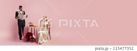 Man in velvet jacket and cravat reads newspaper while woman in satin gown and gloves drinks tea at table against pink studio background 115477352