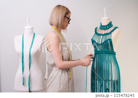 Designer creates unique green macrame dress on dummy in sewing atelier blonde woman ties knot on unfinished garment made of threads in dressmaking workshop Designer creates unique green macrame dress on dummy in sewing atelier blonde woman ties knot on unfinished garment made of threads in dressmaking workshop 115478245
