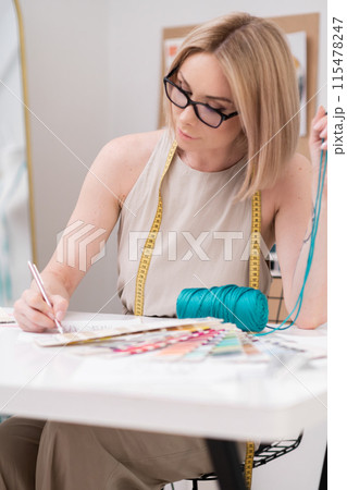 Woman fashion designer draws sketch holding green thread at desk in sewing atelier clothing dressmaker creates elegant outfit models working in tailor studio Woman fashion designer draws sketch holding green thread at desk in sewing atelier clothing dressmaker creates elegant outfit models working in tailor studio 115478247