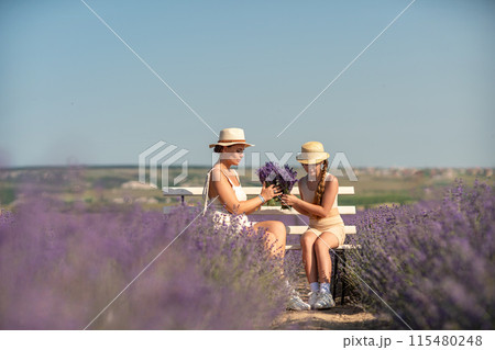 A woman and a child are sitting on a bench in a field of purple flowers lavande. The woman is holding a bouquet of flowers and the child is holding a bouquet of flowers as well A woman and a child are sitting on a bench in a field of purple flowers lavande. The woman is holding a bouquet of flowers and the child is holding a bouquet of flowers as well 115480248
