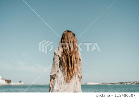 woman with long hair stands in front of a blue sky. She is wearing a white and black striped shirt. 115480307
