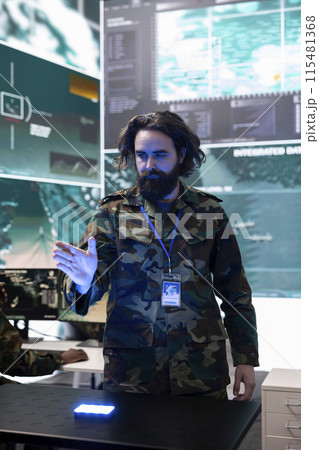 Army soldier examines real time data on a hologram 3d projection, reviewing defense operation details in military control room. Trooper looks at CCTV satellite footage on 3d visualization. 115481368