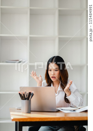 A woman is sitting at a desk with a laptop in front of her. She is looking at the screen with a shocked expression on her face A woman is sitting at a desk with a laptop in front of her. She is looking at the screen with a shocked expression on her face 115484991