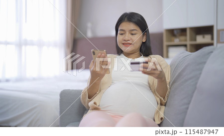 A portrait of happy Asian pregnant woman using a mobile phone device with credit card, shopping online in bedroom at home. Having a baby. Family people lifestyle. Mom love. 115487947