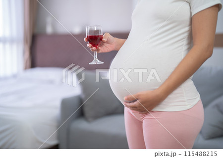 A portrait of happy Asian pregnant woman drinking wine alcohol beverage glass in bedroom at home. Having a baby. Family people lifestyle. Mom. 115488215