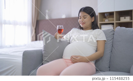 A portrait of happy Asian pregnant woman drinking wine alcohol beverage glass in bedroom at home. Having a baby. Family people lifestyle. Mom. A portrait of happy Asian pregnant woman drinking wine alcohol beverage glass in bedroom at home. Having a baby. Family people lifestyle. Mom. 115488255