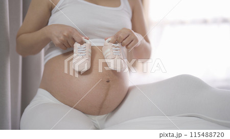 A Portrait of happy Asian pregnant woman with baby shoes in bedroom at home. Having a baby. Family people lifestyle. Love. A Portrait of happy Asian pregnant woman with baby shoes in bedroom at home. Having a baby. Family people lifestyle. Love. 115488720