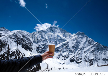 Close-up of a man's hand with a disposable cup against the backdrop of snow-capped mountains and blue sky. Vacation in the mountains, hike, travel. With space for text 115491437
