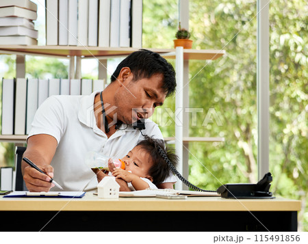 Asian father multitasking at his desk, feeding his baby while talking on telephone, illustrating modern work-life balance in home office 115491856
