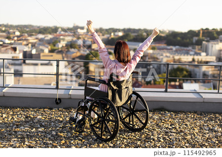 Woman in wheelchair raising arms on rooftop overlooking cityscape Woman in wheelchair raising arms on rooftop overlooking cityscape 115492965