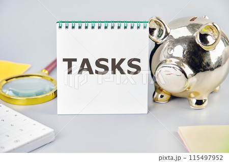 Goals achievement and business success. TASK word on a blank sheet in a notebook in a composition with a piggy bank, magnifying glass, calculator, stickers on a gray background 115497952