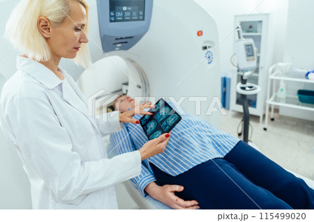 Female patient is lying at CT bed and listening to doctor. Caucasian female doctor looking at tablet with film of MRI tomography scan of the brain. Female patient is lying at CT bed and listening to doctor. Caucasian female doctor looking at tablet with film of MRI tomography scan of the brain. 115499020