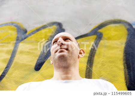 Bald man in white t-shirt looking sideways, colorful graffiti background, urban setting Bald man in white t-shirt looking sideways, colorful graffiti background, urban setting 115499605