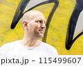 Bald man in white t-shirt looking sideways, colorful graffiti background, urban setting 115499607