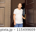 Bald man in white t-shirt holding door handle, looking seriously, outdoor urban setting 115499609