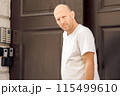 Bald man in white t-shirt holding door handle, standing at entrance, looking forward 115499610