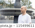 Bald man in white t-shirt standing near fountain, looking thoughtful, outdoor setting 115499611