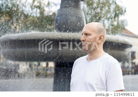 Bald man in white t-shirt standing near fountain, looking thoughtful, outdoor setting 115499611