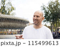 Bald man in white t-shirt smiling, pointing near fountain, outdoor park setting 115499613